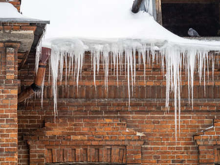 Large icicles hanging from the roof of an old brick building. Roof of old building covered with sharp icicles. Ice stalactite. Poor thermal insulation of the roof leads to the formation of iciclesの写真素材