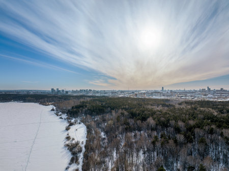 Snow-covered forest on lake shore with ice at sunset and the city on horizon, auerial view. Beautiful winter forest landscape. Lake Shartash and Yekaterinburg, Russiaの写真素材