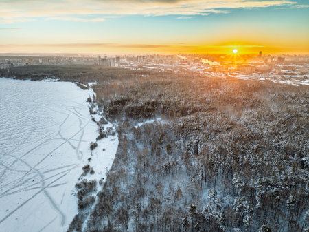 Snow-covered forest on lake shore with ice at sunset and the city on horizon, aerial view. Beautiful winter forest landscape. Lake Shartash and Yekaterinburg, Russiaの写真素材