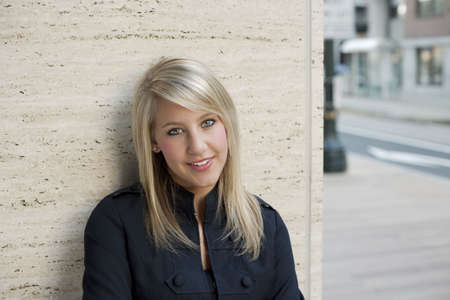 Attractive young woman in a black jacket smiles towards the camera as she leans back against a wall. Horizontal shot.の写真素材