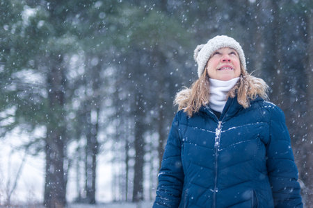 smiling middle age woman looking up at snowflakes falling in forest on hiking trail in winter