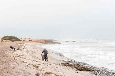 SWAKOPMUND, NAMIBIA - JUNE 30, 2017: A man on a bicycle, a dog and an angler at Swakopmund in the Namib Desert on the Atlantic Coast of Namibiaのeditorial素材