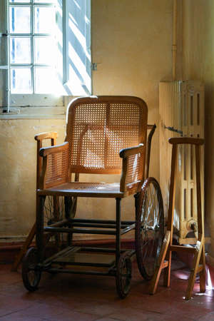 Old wooden wheelchair in the interior. Pale yellow walls, window grille and radiator.の写真素材