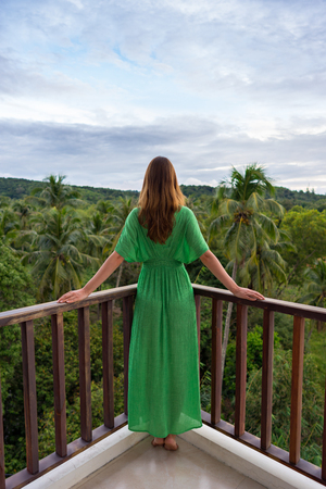A young woman in green dress is standing on a balcony in tropical country and look forward on jungle. Backward view on modelの写真素材