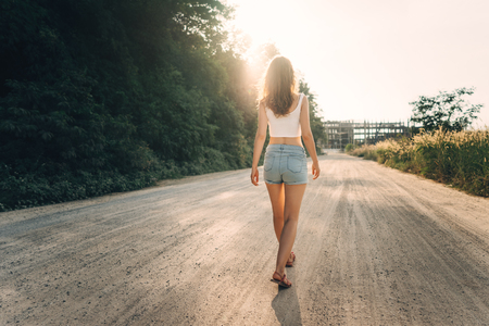 A young slender girl with her hair down goes on the road ahead in the summer direct light. Full-length rear viewの写真素材