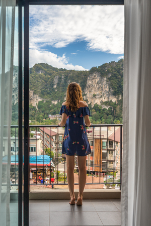 Young woman is standing on balcony in hotel in tropical country and look forward on big cliff and street. Rear view on female.の写真素材