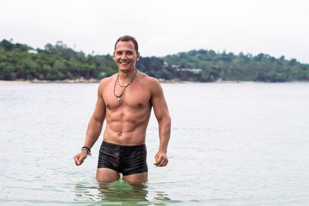 Young strong handsome smiling sporty man with muscle body stands in water in the sea on vacations in Thailandの写真素材