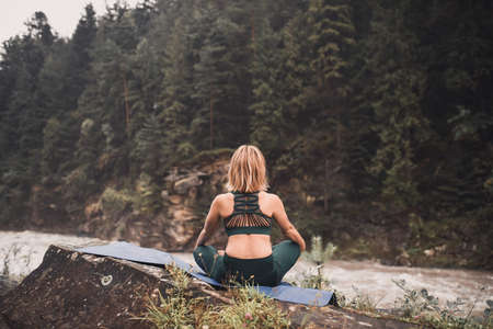 A young blonde woman practices yoga on nature on a stone near a raging river. Green tracksuit.の写真素材