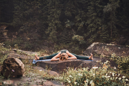 A young blonde woman practices yoga on nature on a stone near a raging river. Green tracksuit.