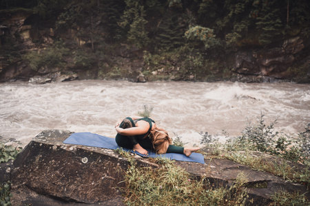 A young blonde woman practices yoga on nature on a stone near a raging river. Green tracksuit.