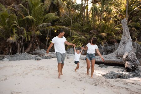 Happy smile european family have rest and run on white sand beach. Father, long black chestnut hair mother, blond baby boy. white cotton clothes. boho dress. t-shorts.Thailand. Aquamarine crystal seaの写真素材