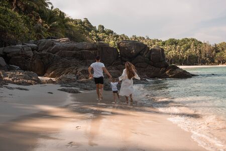 Happy smile european family have rest and run on white sand beach. Father, long black chestnut hair mother, blond baby boy. white cotton clothes. boho dress. t-shorts.Thailand. Aquamarine crystal seaの写真素材