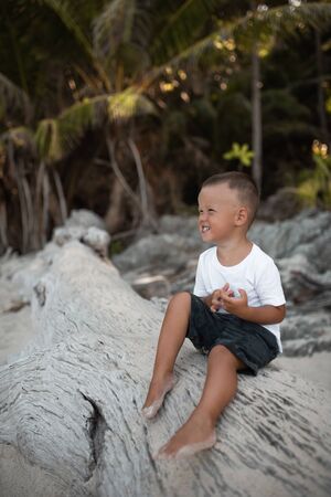 Happy smile european blond baby boy have rest and sit on white sand beach. . white cotton clothes. t-short.Thailand. palms. rocksの写真素材