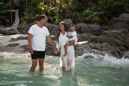Happy smile european family have rest and run on white sand beach. Father, long black chestnut hair mother, blond baby boy. white cotton clothes. boho dress. t-shorts.Thailand. Aquamarine crystal seaの写真素材
