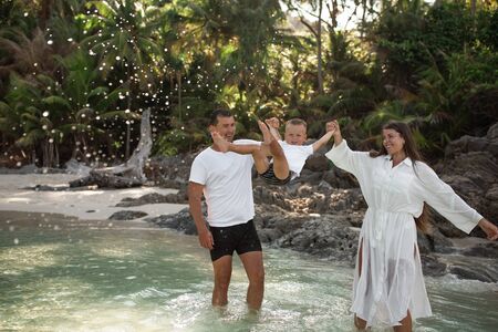 Happy smile european family have rest and run on white sand beach. Father, long black chestnut hair mother, blond baby boy. white cotton clothes. boho dress. t-shorts.Thailand. Aquamarine crystal seaの写真素材