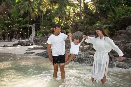 Happy smile european family have rest and run on white sand beach. Father, long black chestnut hair mother, blond baby boy. white cotton clothes. boho dress. t-shorts.Thailand. Aquamarine crystal seaの写真素材