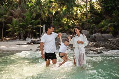 Happy smile european family have rest and run on white sand beach. Father, long black chestnut hair mother, blond baby boy. white cotton clothes. boho dress. t-shorts.Thailand. Aquamarine crystal seaの写真素材