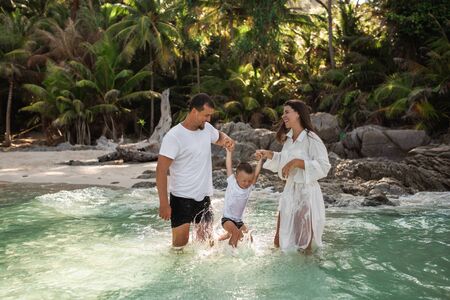 Happy smile european family have rest and run on white sand beach. Father, long black chestnut hair mother, blond baby boy. white cotton clothes. boho dress. t-shorts.Thailand. Aquamarine crystal seaの写真素材