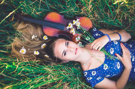 Young slim fair-skinned girl blonde beautiful lies in tall grass. Top view. Girl in a blue short tight dress with a print of daisies. A bouquet and lonely daisies scattered around.A woman hugs violin.の写真素材