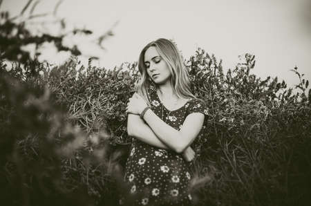 A young blonde European thin girl stands in the midst of a very tall purple and poppy plant hugs herself with her eyes closed. Womens blue short skin-tight dress with white daisy print.の写真素材