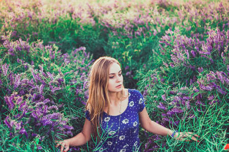 light-skinned European blonde young slim girl walks on tall grass overgrown with purple flowers. Womens blue short skin-tight dress with white daisy print.の写真素材