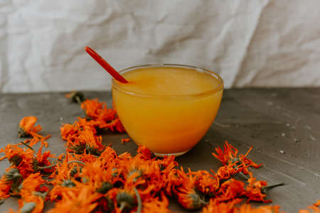 A small cup is filled to the top with yellow liquid honey. Medicinal herbal dried plants marigold, orange calendula. Neutral white and gray background.の写真素材