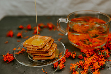 The honey flows in a thin stream onto the baked goods. Orange calendula flower tea in a transparent...