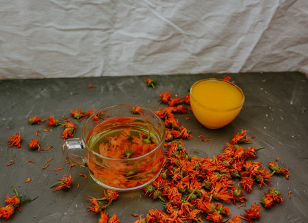 Flower tea in a large transparent mug. A small cup is filled to the top with yellow liquid honey. Medicinal herbal dried plants marigold, orange calendula. Neutral white and gray background.の写真素材