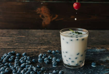 Blueberries float in a transparent glass with milk. Green basil leaves in a glass. a womans hand holds a cherry and soaks it in milk. Blue fresh blueberries are scattered on an old brown wooden tableの写真素材