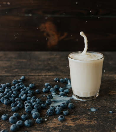 Berries are thrown into the transparent glass making splashes of milk. Blue fresh blueberries are scattered on an old brown wooden cracked table. White water pour down on the table.の写真素材