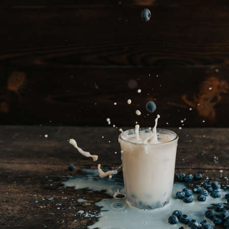 Berries are thrown into the transparent glass making splashes of milk. Blue fresh blueberries are scattered on an old brown wooden cracked table. White water pour down on the table.の写真素材