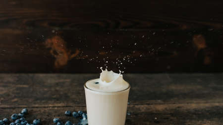 Berries are thrown into the transparent glass making splashes of milk. Blue fresh blueberries are scattered on an old brown wooden cracked table. White water pour down on the table.の写真素材