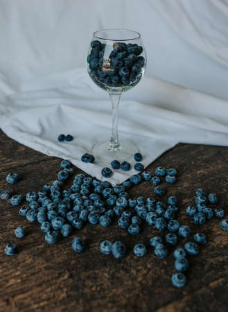 Fresh blue matte round blueberries filled in a transparent glass stands on a wooden brown shabby textured table and berries are scattered everywhereの写真素材