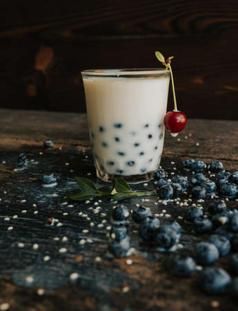 Red cherry hangs on a glass, everything is covered with sesame seeds on top, milk is spilled on the table. Blueberries float in a transparent glass with milk. Blue fresh blueberries are scatteredの写真素材