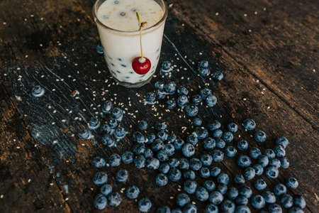Red cherry hangs on a glass, everything is covered with sesame seeds on top, milk is spilled on the table. Blueberries float in a transparent glass with milk. Blue fresh blueberries are scatteredの写真素材