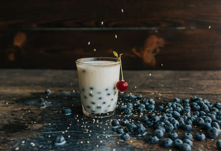 Red cherry hangs on a glass, everything is covered with sesame seeds on top, milk is spilled on the table. Blueberries float in a transparent glass with milk. Blue fresh blueberries are scatteredの写真素材
