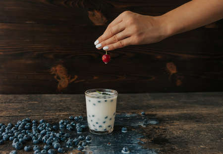 Blueberries float in a transparent glass with milk. Green basil leaves in a glass. a womans hand holds a cherry and soaks it in milk. Blue fresh blueberries are scattered on an old brown wooden tableの写真素材