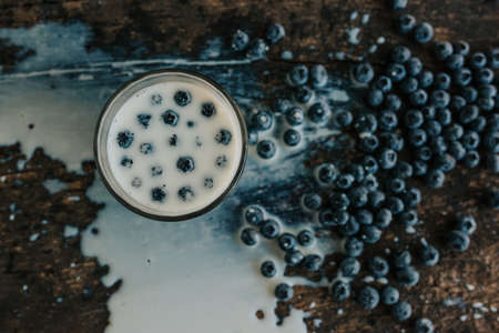Berries are thrown into the transparent glass making splashes of milk. Blue fresh blueberries are scattered on an old brown wooden cracked table. White water pour down on the table.の写真素材
