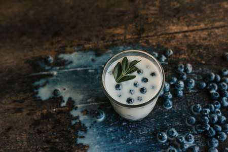 Blueberries float in a transparent glass with milk. Green basil leaves in a glass. Blue fresh blueberries are scattered on an old brown wooden cracked table. White water spilled.の写真素材
