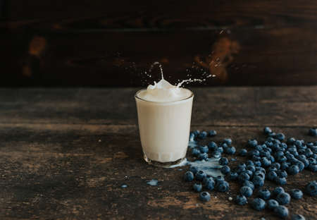 Berries are thrown into the transparent glass making splashes of milk. Blue fresh blueberries are scattered on an old brown wooden cracked table. White water pour down on the table.の写真素材