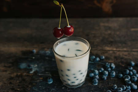 Blueberries float in a transparent glass with milk. Green basil leaves in a glass. a womans hand holds a cherry and soaks it in milk. Blue fresh blueberries are scattered on an old brown wooden tableの写真素材