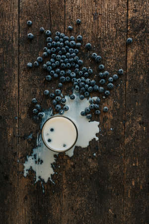 Berries are thrown into the transparent glass making splashes of milk. Blue fresh blueberries are scattered on an old brown wooden cracked table. White water pour down on the table.の写真素材