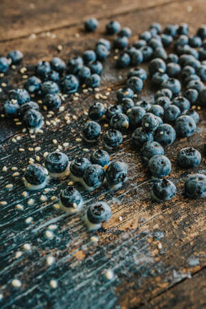 Blue fresh blueberries are scattered on an old brown wooden cracked table. Sesame seeds were poured between the fruits. Wet drops of milk on the table. basil leaves, green fresh seasoning.の写真素材