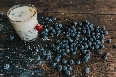 Red cherry hangs on a glass, everything is covered with sesame seeds on top, milk is spilled on the table. Blueberries float in a transparent glass with milk. Blue fresh blueberries are scatteredの写真素材