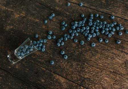 Fresh blue matte round blueberries filled in a transparent glass stands on a wooden brown shabby textured table and berries are scattered everywhereの写真素材