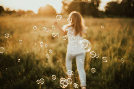 a little blonde girl dressed in light-colored clothes and a white T-shirt runs away from soap bubble. Along the tall green grass towards the setting sun. Warm summer sunny weather at sunset.の写真素材