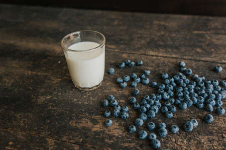 A transparent glass with milk. Blue fresh blueberries are scattered on an old brown wooden cracked table.の写真素材