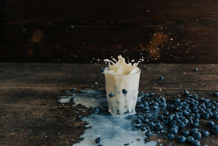Berries are thrown into the transparent glass making splashes of milk. Blue fresh blueberries are scattered on an old brown wooden cracked table. White water pour down on the table.の写真素材