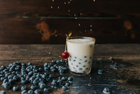 Red cherry hangs on a glass, everything is covered with sesame seeds on top, milk is spilled on the table. Blueberries float in a transparent glass with milk. Blue fresh blueberries are scatteredの写真素材