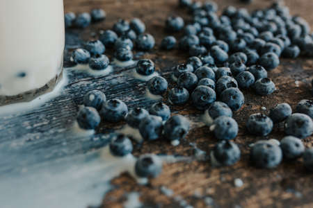 Berries are thrown into the transparent glass making splashes of milk. Blue fresh blueberries are scattered on an old brown wooden cracked table. White water pour down on the table.の写真素材